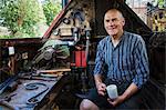 Blacksmith sitting on a working boat, a narrowboat on a waterway, holding a mug, smiling at camera.