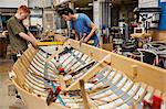 Two men in a boat-builder's workshop, working together on a wooden boat hull.