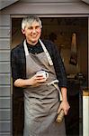 A craftsman wearing a work apron standing in doorway of a workshop, holding mug and small log of wood, smiling. Tea break.