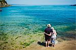 Rear view of elderly couple standing with arms around each other looking out over the vivid blue and turquoise coloured water, the ocean.