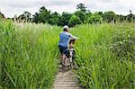 Rear view of woman pushing bicycle along path through tall grass.