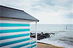 A blue and white striped painted beach hut on a beach on the coast.