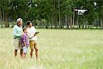 Man flying remote control drone in open field while older man and boy watch