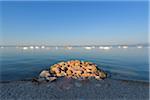 Sunlit rocks piled on beach at shoreline of Lake Garda (Lago di Garda) with boats anchored in the distance on a sunny morning at Bardolino in Veneto, Italy