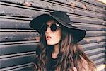 Portrait of young woman standing beside shutter, wearing floppy hat and sunglasses