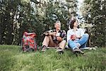 Mature couple relaxing on grass, holding tin cups, rucksack beside them