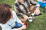 Mature couple crouching on grass, man pouring hot drink from drinks flask, elevated view