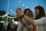 Two mature female friends taking smartphone selfie at night market festival in park, London, UK