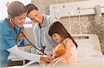 Female nurse using stethoscope on teddy bear of girl patient in hospital room