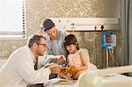 Male doctor using otoscope on teddy bear of girl patient in hospital room