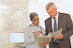 Female doctor and male hospital administrator talking, looking at digital tablet and paperwork in examination room