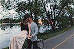 Romantic young man whispering to girlfriend on bicycle handlebars by lake at dusk