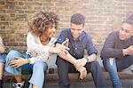 Four friends sitting in street, laughing, young woman holding smartphone