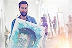 Portrait smiling, confident, proud male artist holding painting in art class studio