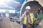 Male engineer with laptop talking on cell phone in steel factory