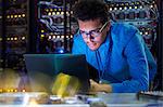 Focused male IT technician working at laptop in dark server room