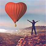 Girl with hands up stand on top of mountain looking at big red air balloon heart shape flying in blue sky. Romantic trip on Valentine's Day. Sports, travel and recreation. Nature background. Back view