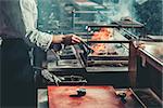 Food concept. Chef in white uniform monitors the degree of roasting and greases with a brush meat with oil sause in interior of restaurant kitchen. Preparing traditional beef steak on barbecue oven.