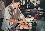 One young white chef dressed in white uniform decorate ready dish in restaurant. He is working on maki rolls. Preparing traditional japanese sushi set in interior of modern professional kitchen