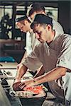 Three young chefs dressed in white uniform decorate ready dish in restaurant. They are working on maki rolls. Preparing traditional japanese sushi set in interior of modern professional kitchen