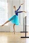 Attractive ballerina stands on the left toe next to the ballet barre on the white wall and windows background. She wears a lace blue leotard, cyan tutu, pointe shoes and looks into camera. Indoors.