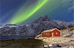 Northern Lights illuminate snowy peaks and the wooden cabin on a starry night at Budalen Svolvaer Lofoten Islands Norway Europe