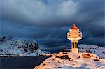 A lighthouse in the snow in the Arctic night with the village of Reine in the background Nordland Lofoten Islands Norway Europe