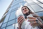 Businesswoman outside office building, holding coffee cup and smartphone, tattoos on hands, low angle view