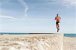 Young woman running along sea wall, mid air