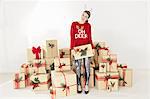 Young woman in red sweater and reindeer antlers by stacked christmas gifts holding christmas parcel