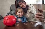 Mature woman taking smartphone selfie with daughter and piggy bank on coffee table