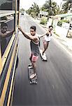 Two young people skateboarding while holding on to a moving school bus.