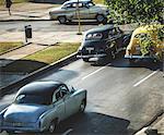 Classic 1950s cars driving on a road at a junction.