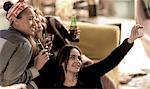 Four young women sitting on a sofa, laughing, taking a selfie, holding beer bottles.