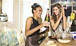 Two young women at a party in sequined dresses drinking and laughing, one holding a champagne bottle.