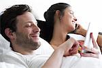 Couple relaxing in bed, man using digital tablet while woman makes phone call