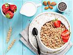 Top view of bowl with muesli, fresh strowberry, nuts, chia seeds and milk in milk jug on blue wooden background. Copy space.