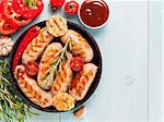 Top view of chicken homemade sausages, sauces ketchup and vegetables and herbs on blue wooden background. Grilled sausages and grilled vegetables in black iron pan. Copy space. Top view or flat lay.
