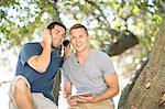 Two young male friends sitting under tree listening to headphones