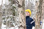 Boy in yellow knit hat looking up at tree in snow covered forest