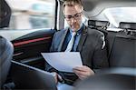 Businessman sitting in back of car, using laptop, holding document