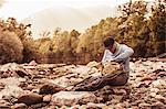 Fisherman sitting on river rocks searching fishing bag, Mozirje, Brezovica, Slovenia