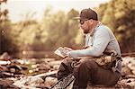 Fisherman sitting on river rocks looking at smartphone, Mozirje, Brezovica, Slovenia