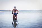 Pensive, determined female open water swimmer with hands on hips in ocean surf