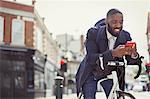 Young businessman commuting with bicycle, texting with cell phone on sunny urban street