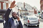 Young businessman talking on cell phone and hailing taxi on sunny urban street