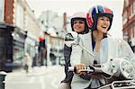 Smiling young women friends wearing helmets and riding motor scooter on urban street