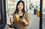 Young woman with coffee texting with cell phone on urban sidewalk