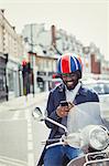 Smiling young businessman in helmet on motor scooter, texting with cell phone on urban street