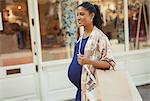 Smiling pregnant woman with shopping bags walking along storefront